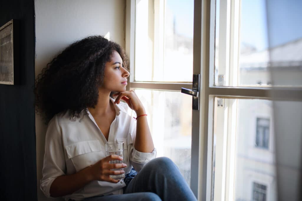 Portrait of young woman sitting on windowsill indoors, looking out and daydreaming