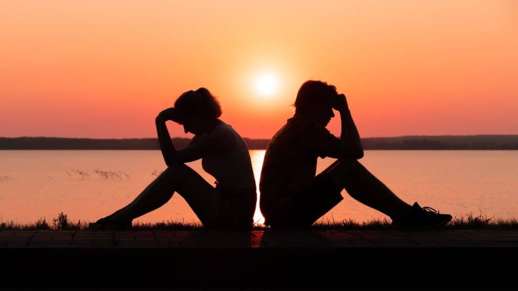 Silhouette of an upset couple sitting on the shore of the lake at sunset. Relationship problems